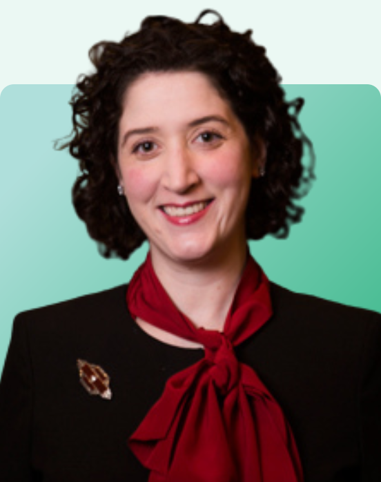 A woman with short curly dark hair, wearing a black blazer with a brooch and a red scarf tied at the neck, smiles in front of a light green gradient background.