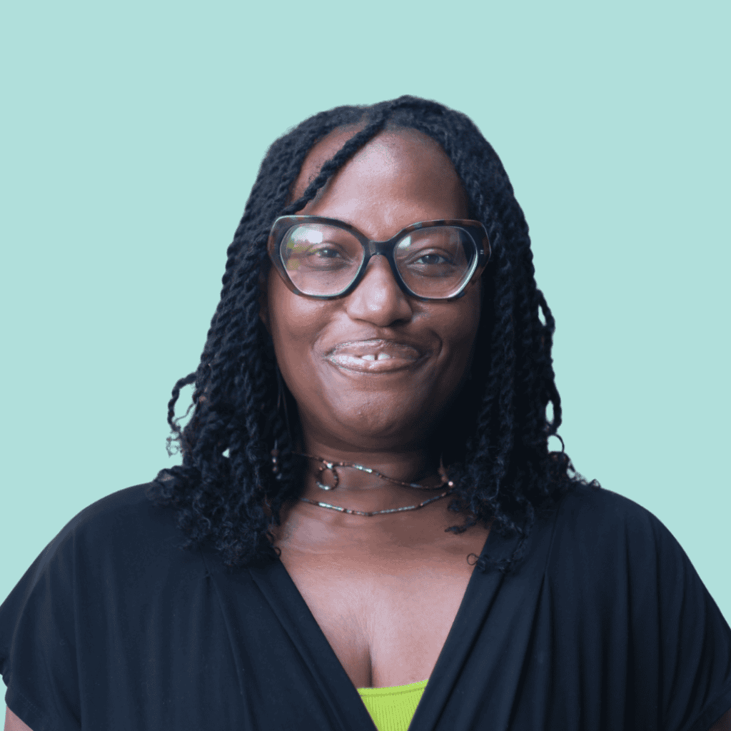 A woman with shoulder-length curly hair and glasses smiles at the camera. She is wearing a black top over a green shirt and a layered necklace, with a light blue background.