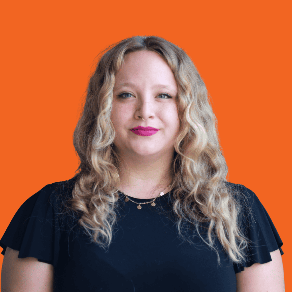 A woman with wavy blonde hair and light skin, wearing a black top and a necklace, stands in front of an orange background, looking at the camera and smiling slightly.