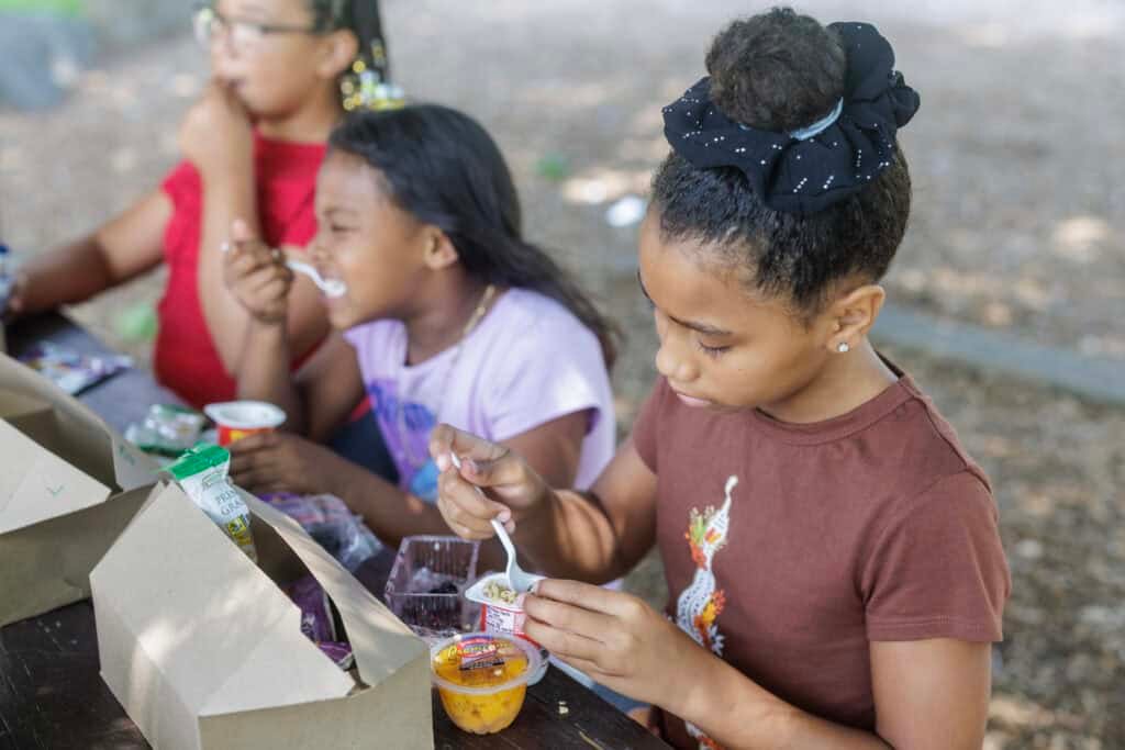 Three young girls sit at a picnic table outdoors, eating snacks from cardboard lunch trays. The girl in the foreground is focused on her food, holding a spoon and looking down at a cup of fruit.