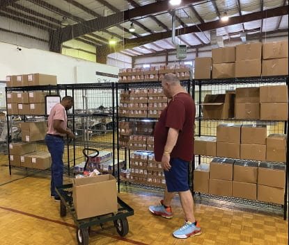 Two men stand next to metal shelves stacked with cardboard boxes in a warehouse. One man loads boxes onto a cart, while the other observes. The floor is wooden and the ceiling is industrial.