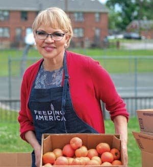 A woman wearing glasses and a Feeding America apron smiles while holding a box of tomatoes outdoors, with buildings and grass visible in the background.