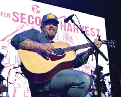 A man wearing a blue shirt and cap plays an acoustic guitar and sings into a microphone on stage. A large sign behind him reads SECOND HARVEST.
