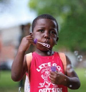 A young child with dark skin wears a red sleeveless shirt and backpack, blowing soap bubbles outdoors. The background is blurred with greenery and a building visible.