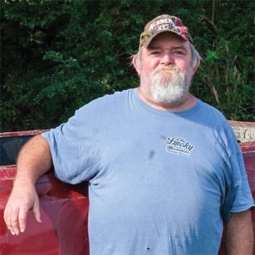 A bearded man in a blue t-shirt and camo cap leans on a red pickup truck, smiling, with green foliage in the background.