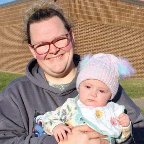 A smiling adult with glasses holds a baby wearing a colorful knit hat and patterned outfit. They are outside on a sunny day, standing in front of a brick building.
