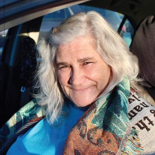 Older woman with long white hair sits in a car, smiling warmly at the camera. She wears a blue top and a colorful patterned scarf draped over her shoulders. Sunlight illuminates her face.