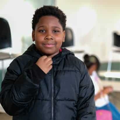 A young boy wearing a black jacket stands indoors, looking at the camera and touching his collar. In the blurred background, another child is seated on the floor.