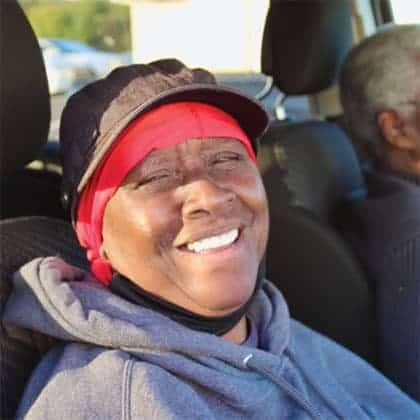 A woman wearing a red headscarf, black cap, and gray hoodie smiles while sitting in a car. Sunlight is shining on her face, and another person is seated beside her in the background.