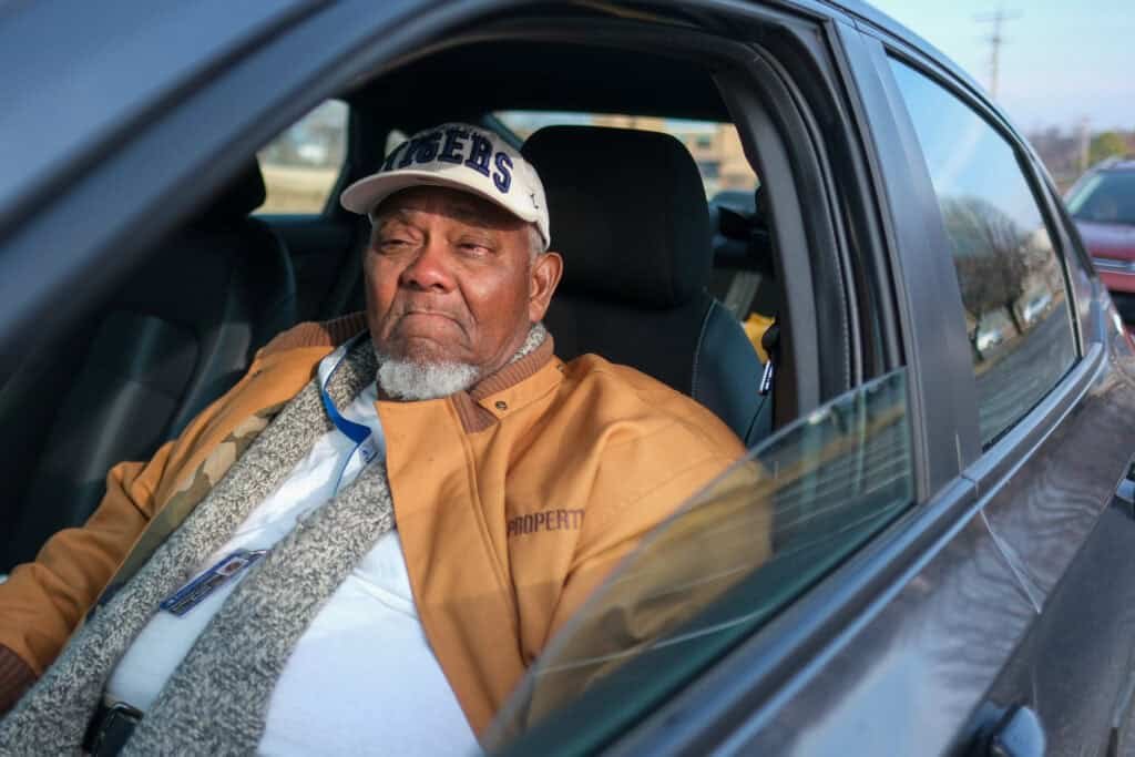 An elderly man in Nashville sits in the driver’s seat of his car after receiving food assistance, wearing a tan jacket and white cap as sunlight fills the car.