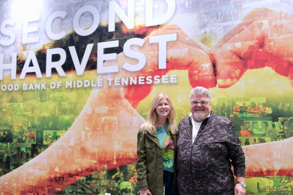 A smiling man and woman stand in front of a colorful mural with the words Second Harvest Food Bank of Middle Tennessee in large letters, capturing the heart of Nashville’s country music spirit.