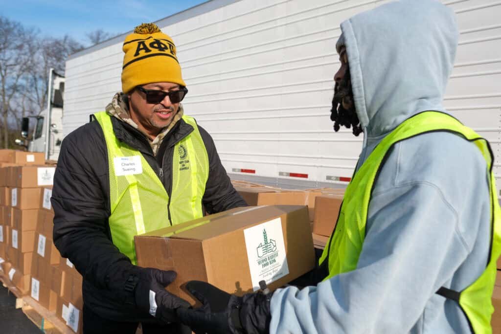 Two volunteers wearing safety vests hand off a box during a holiday food distribution event in Nashville, standing beside stacked boxes and a large truck. One person wears a yellow winter hat and sunglasses.