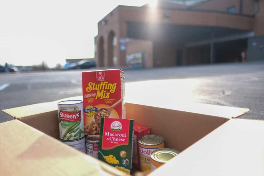 A cardboard box filled with canned food, stuffing mix, and macaroni & cheese sits in a parking lot outside a building on a sunny day.