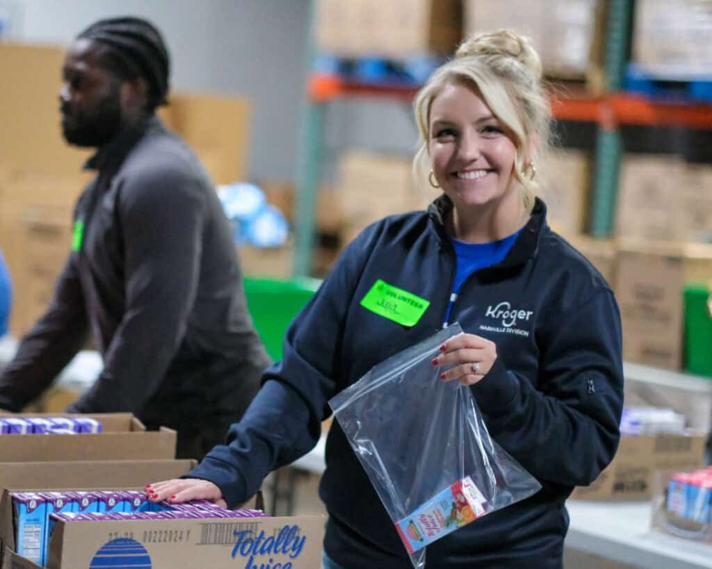 A smiling woman wearing a Kroger jacket and name tag packs food items into a plastic bag at a warehouse, supporting holiday hunger relief in Middle Tennessee. Another person works in the background among boxes on a table.