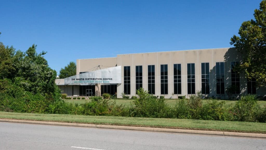 A beige, two-story building with tall windows and a sign reading The MATT Distribution Center stands behind green shrubs and grass on a sunny day, offering support for those facing post-holiday debt and food insecurity. A sidewalk and road run in front of the building.
