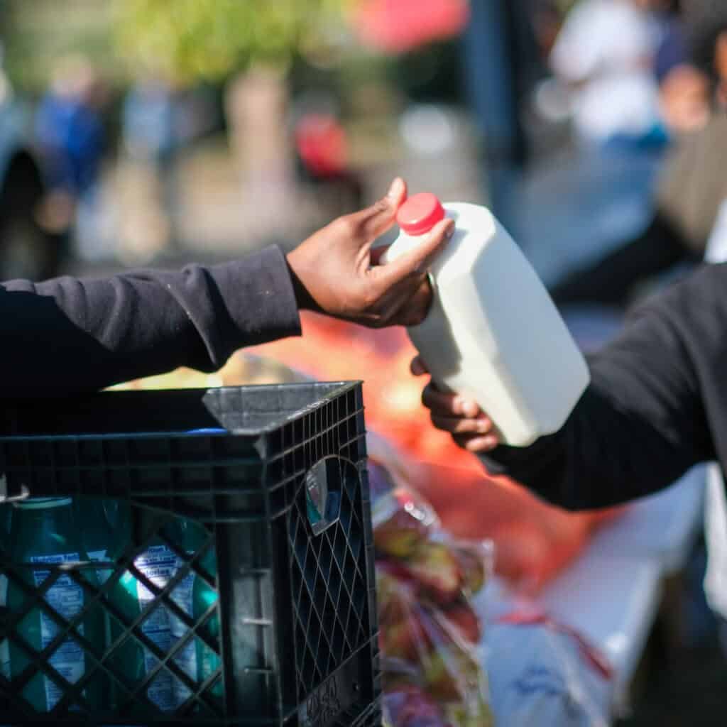 At an outdoor food distribution event, a person hands a bottle of milk to another as seasonal jobs ending in January create increased demand. A crate of supplies and bags of apples are visible in the foreground.