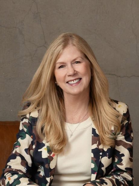 A woman with long blonde hair smiles at the camera. She is wearing a patterned blazer over a light top and is seated in front of a gray, textured wall.