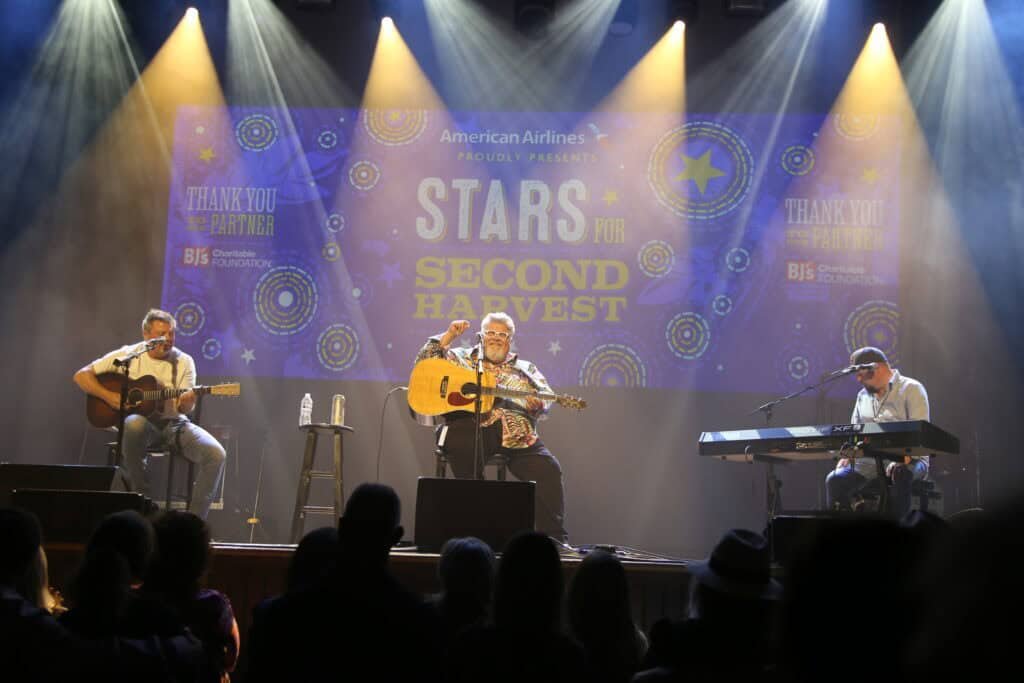 Three musicians perform Nashville Country Music on stage with guitars and a keyboard at an event called “Stars for Second Harvest,” as projected on the screen behind them, while an audience watches in the foreground.