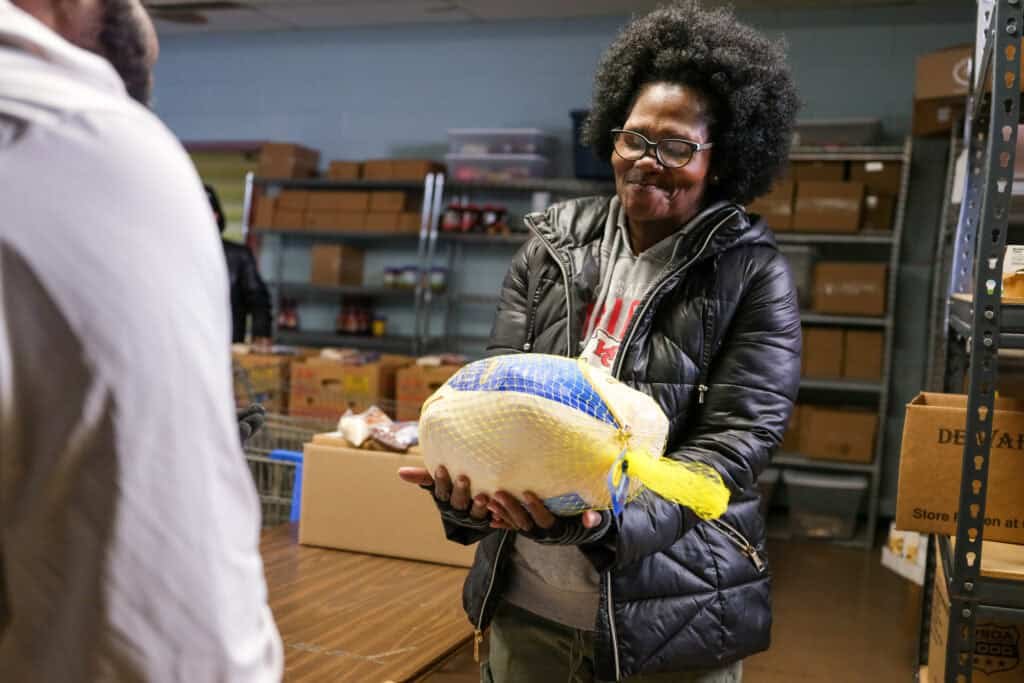 A woman wearing glasses and a black jacket smiles while holding a packaged turkey in a food pantry, with shelves of boxes and canned goods visible in the background.