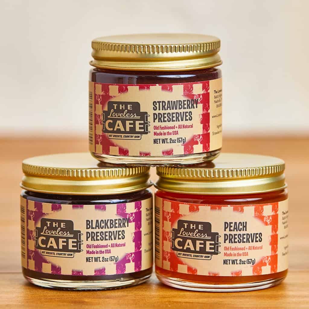 Three small jars of preserves are stacked on a wooden surface. The flavors are strawberry, blackberry, and peach, each labeled with colorful, patterned designs and gold lids. The brand The Loveless Café is visible on each jar.
