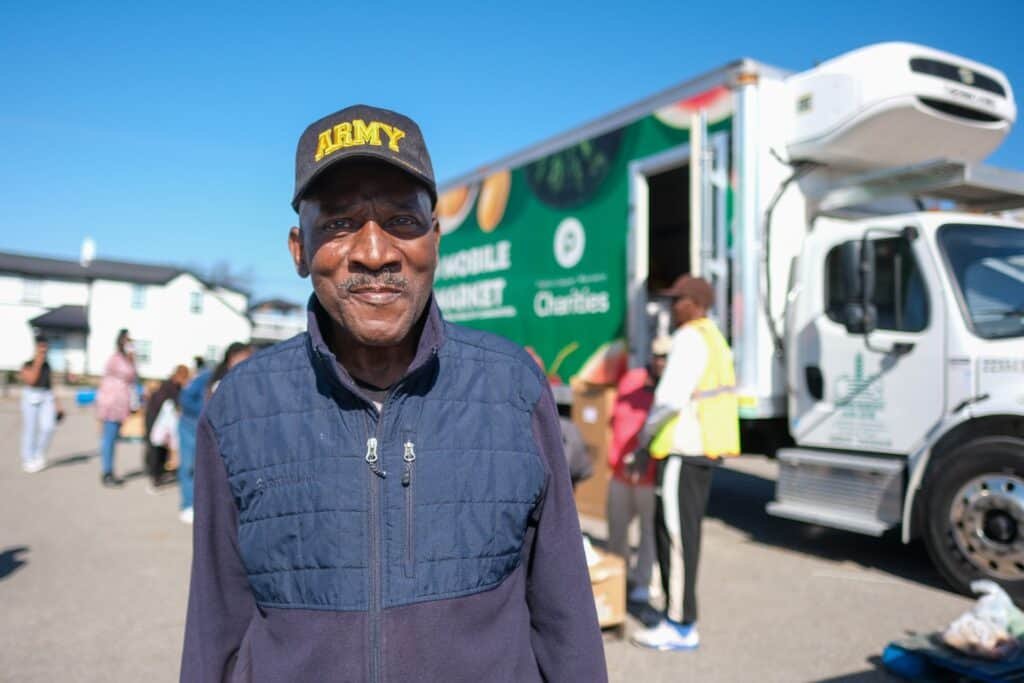 A man wearing an ARMY cap and blue vest smiles at the camera in front of a mobile food pantry truck, with volunteers and people in the background on a sunny day.