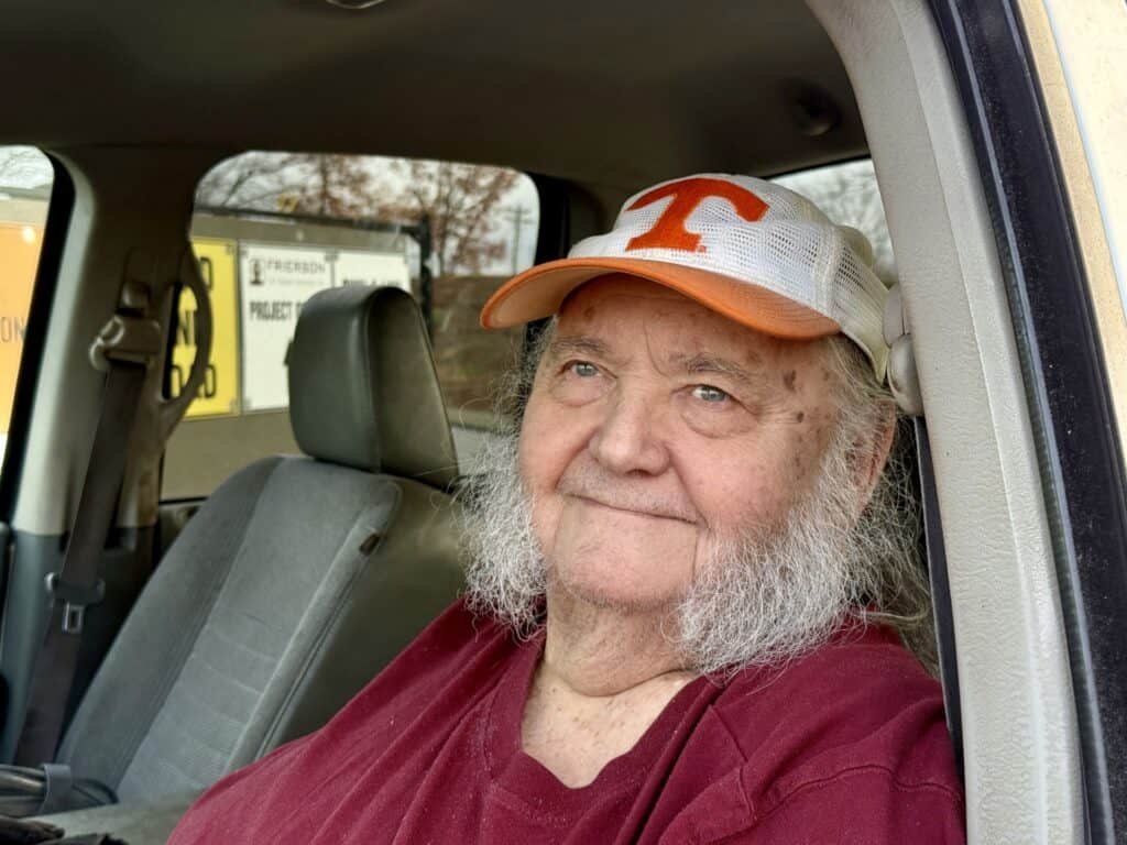 An older person with long white sideburns and a maroon shirt smiles while sitting in a car. They are wearing a white and orange hat with a T on it. The car door is open and the background shows a building and trees.