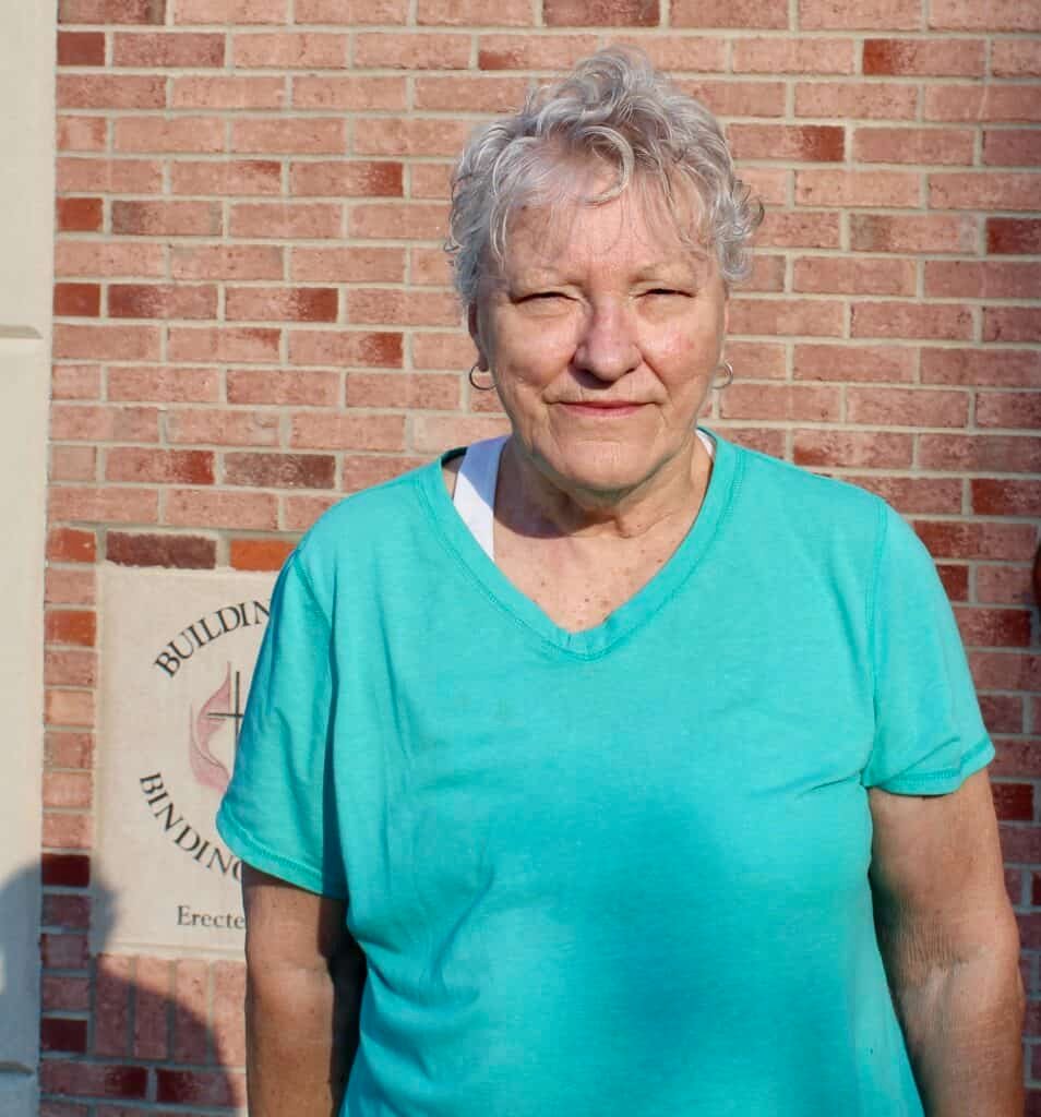 An older woman with short, curly gray hair wearing a teal shirt stands in sunlight in front of a red brick wall, with part of a building plaque visible in the background.