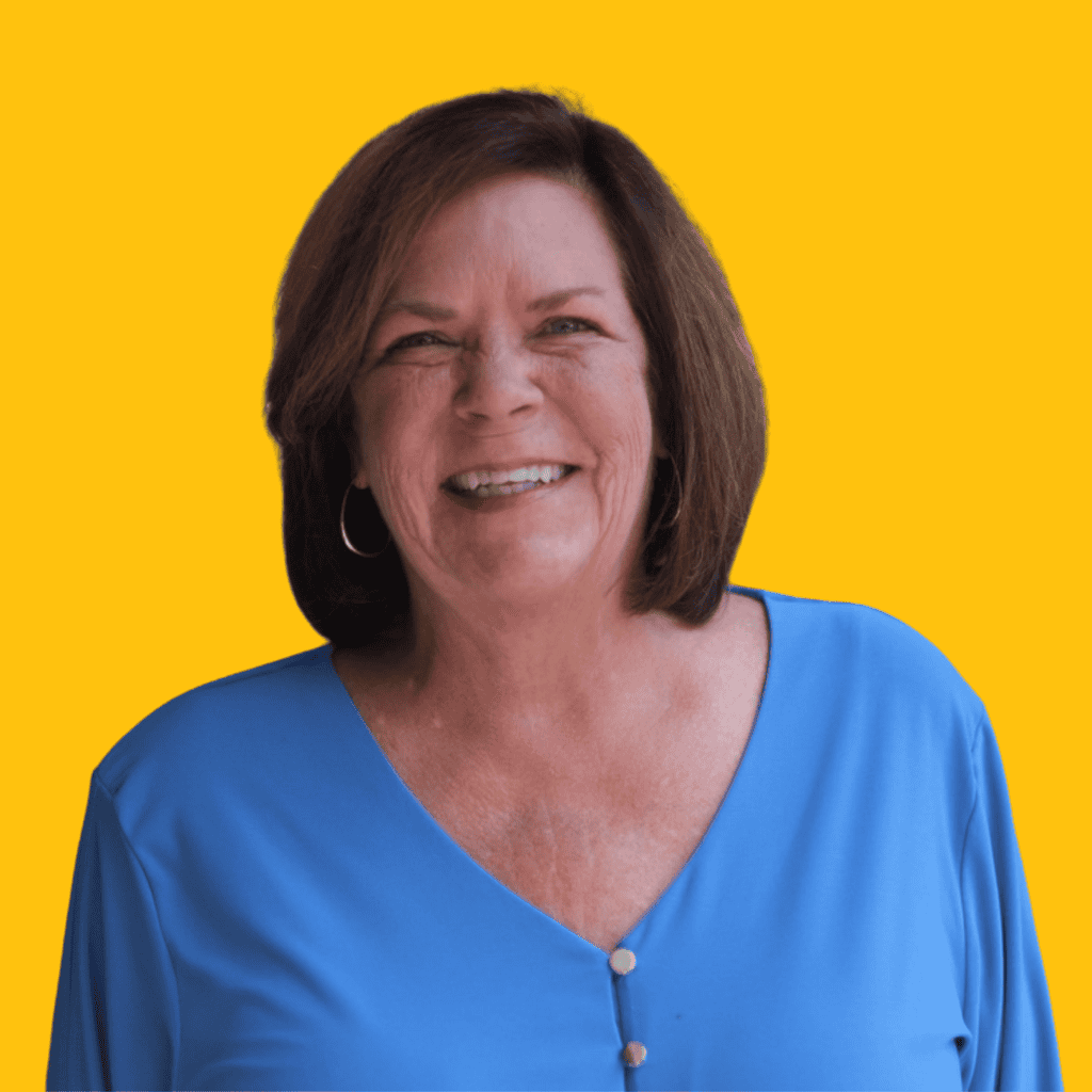 A smiling middle-aged woman with short brown hair, wearing a light blue blouse with buttons, stands against a bright yellow background.