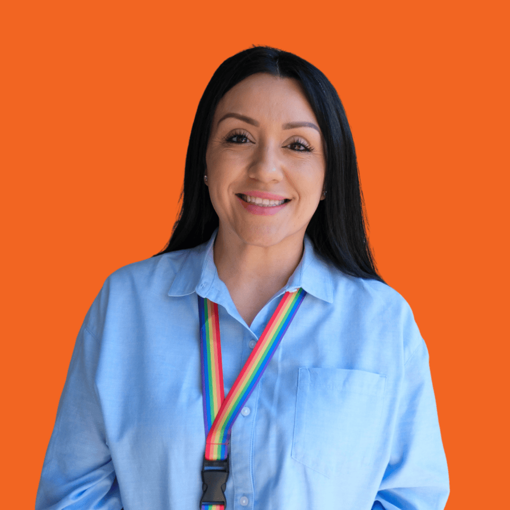 A woman with long dark hair smiles at the camera. She is wearing a light blue shirt and a rainbow lanyard, standing in front of a bright orange background.