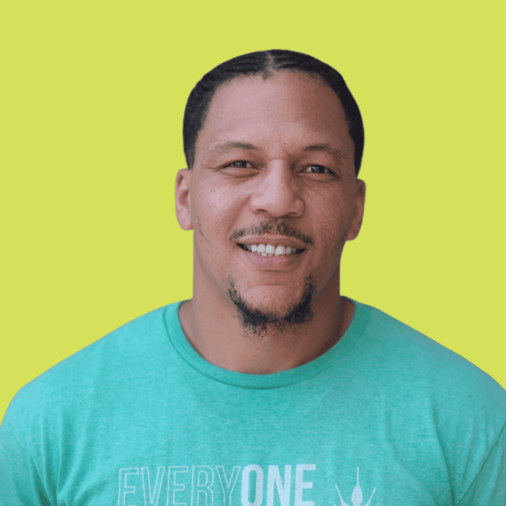 A smiling man with short hair and a goatee wearing a teal shirt that reads EVERYONE stands against a bright yellow background.