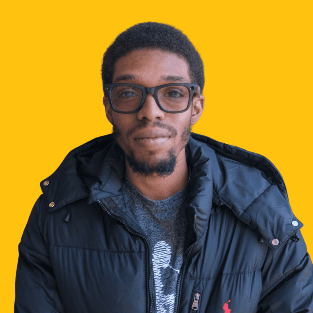 A man with short hair and a beard wearing black glasses, a gray shirt, and a black puffer jacket stands in front of a solid yellow background.