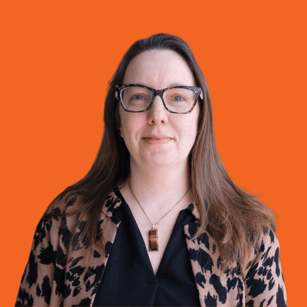 A woman with long brown hair and glasses, wearing a black top, a leopard-print cardigan, and a rectangular pendant necklace, stands in front of an orange background.